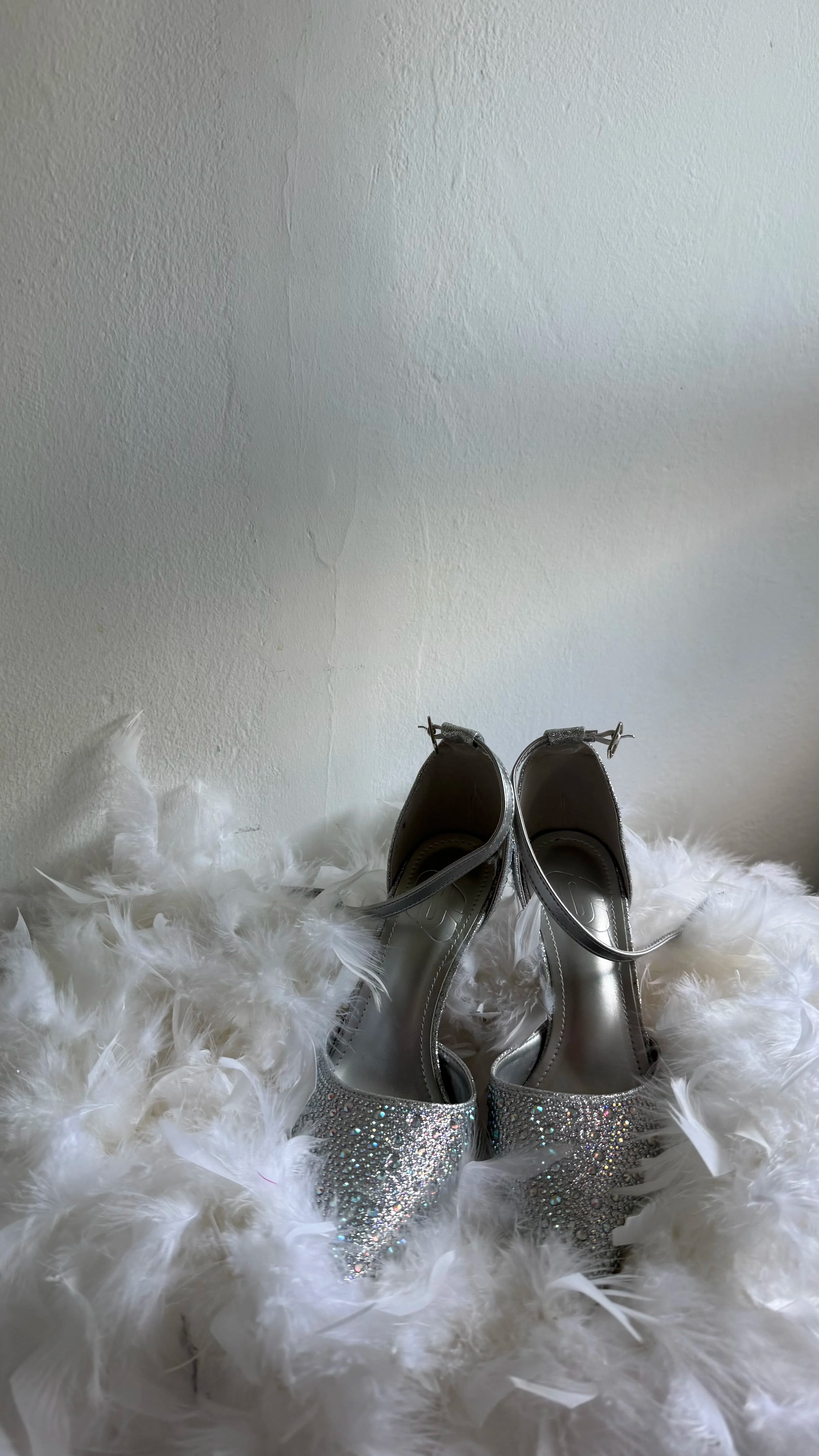 Sandalias de tacón cubiertas de pedrería reposando sobre un lecho de plumas blancas, junto a una pared encalada.