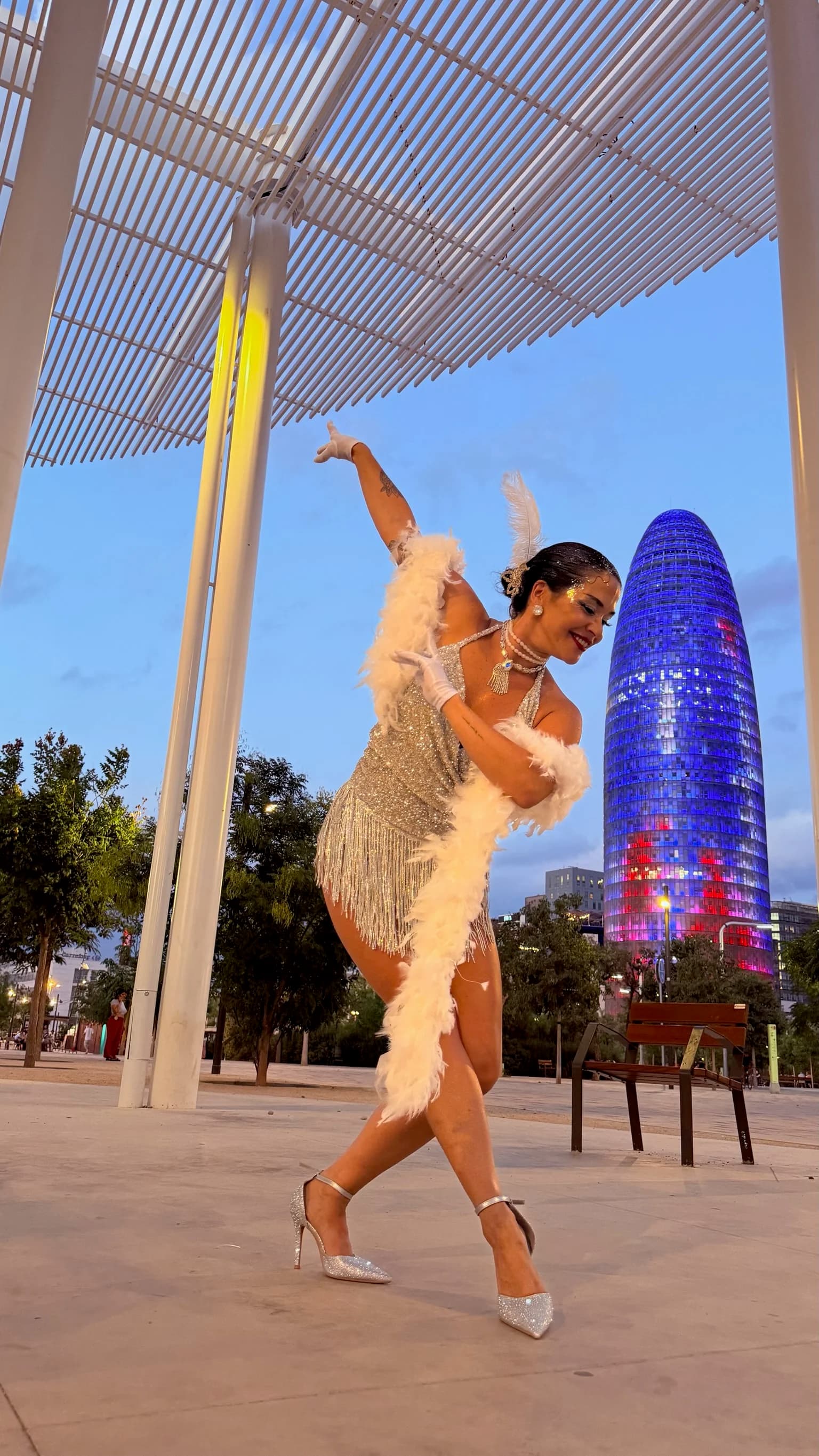 Bailarina de Las Coquettes apoyada en una columna, con vestido de lentejuelas y flecos plateados, guantes largos hasta el codo, boa de plumas blancas y sandalias de pedrería.