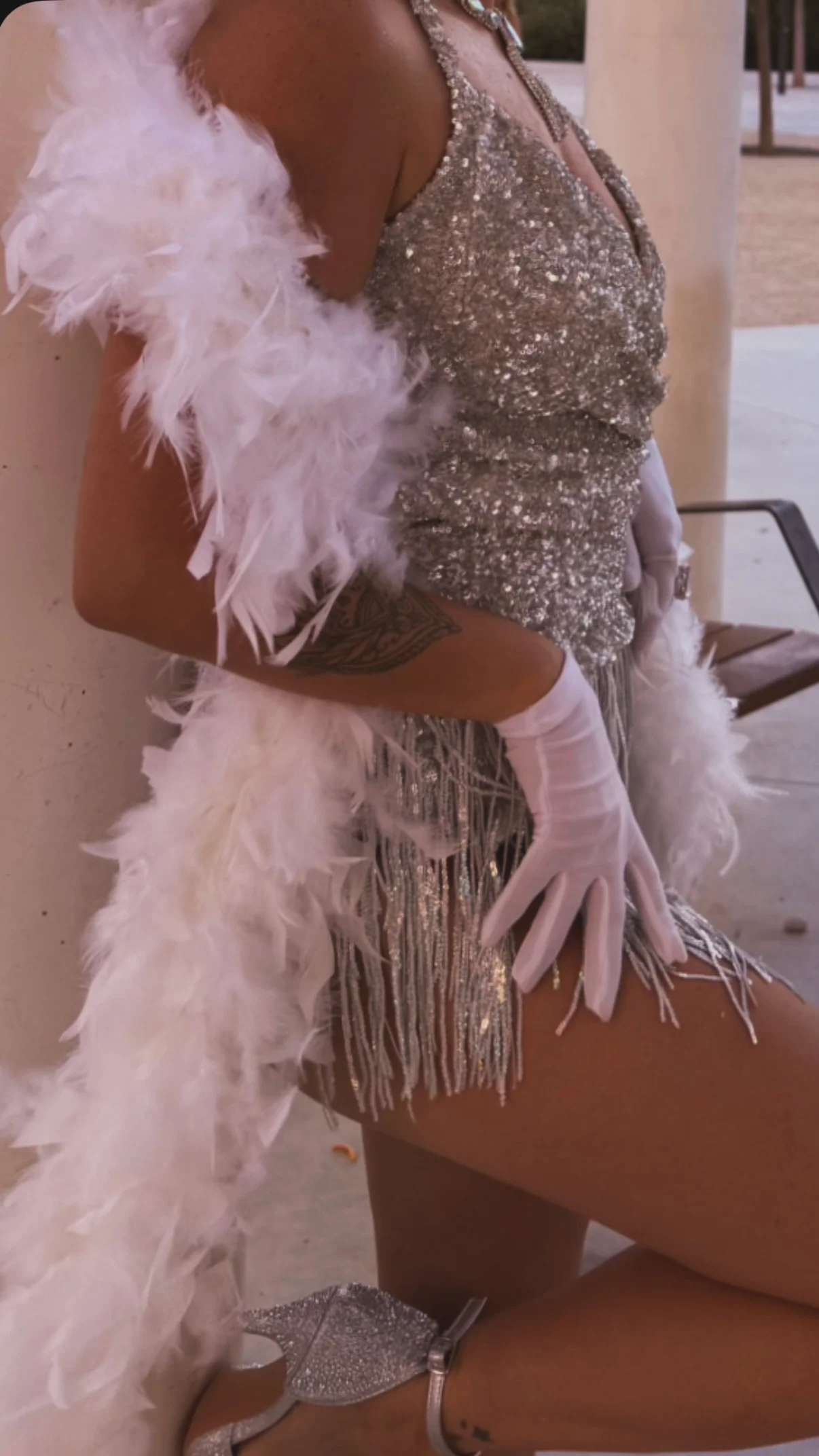 Bailarina de Las Coquettes apoyada en una columna, con vestido de lentejuelas y flecos plateados, guantes largos hasta el codo, boa de plumas blancas y sandalias de pedrería.