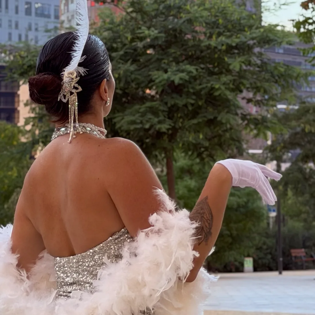 Vista de espalda de una bailarina con moño alto, tocado de pluma y boa de plumas blancas sobre el vestido de lentejuelas.